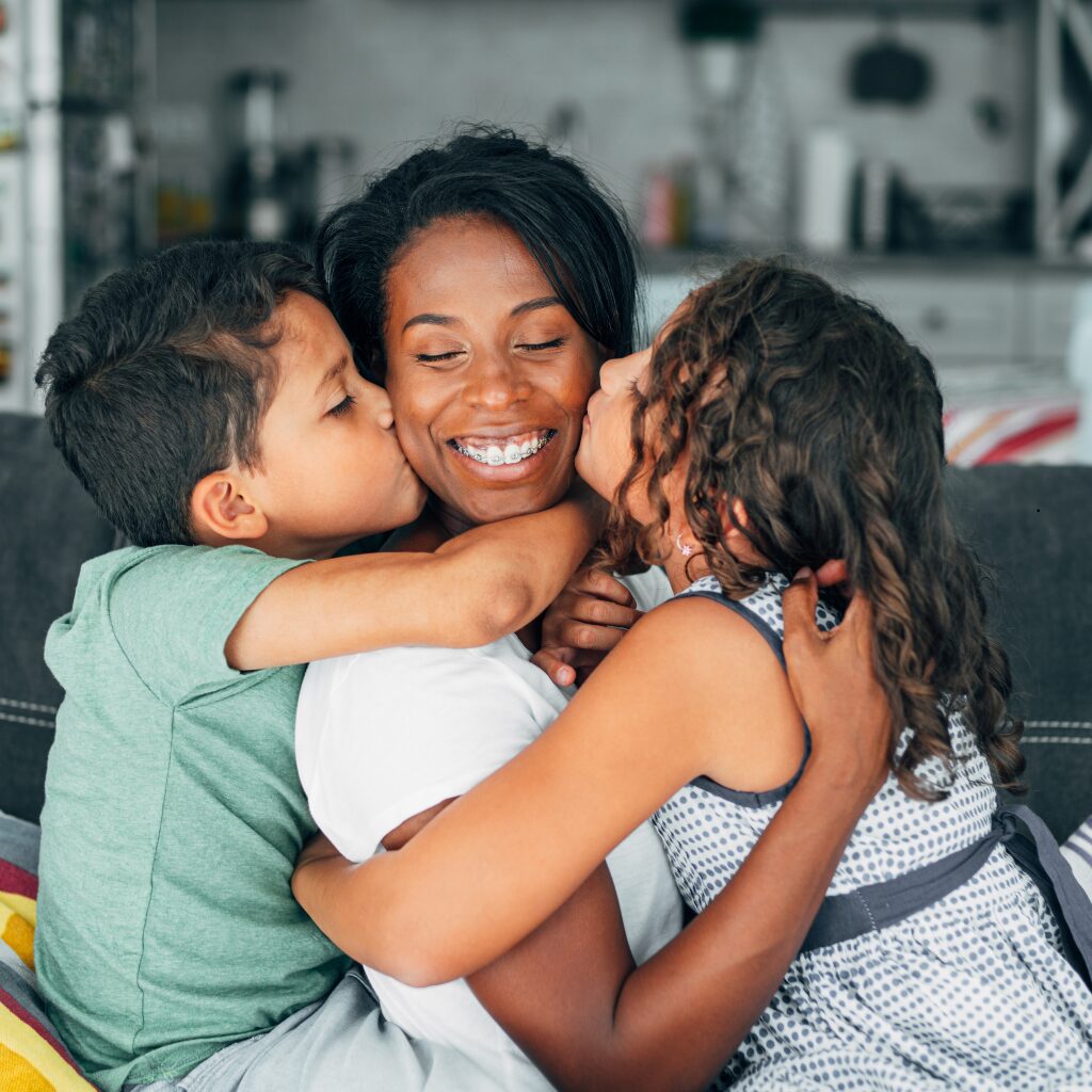 A smiling mother sits on a couch, embraced by her two children who kiss her cheeks at the same time.
