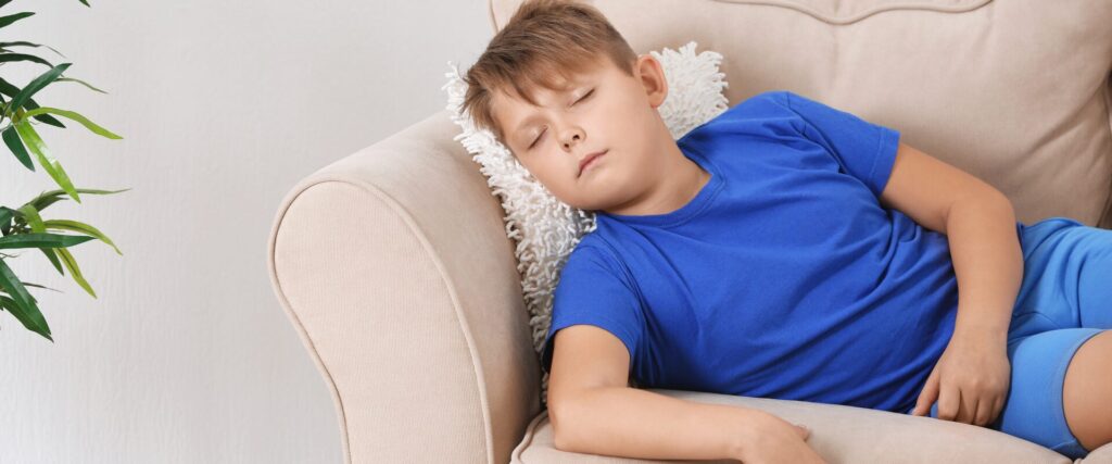 A child wearing a blue shirt and shorts lies on a couch appearing exhausted. 