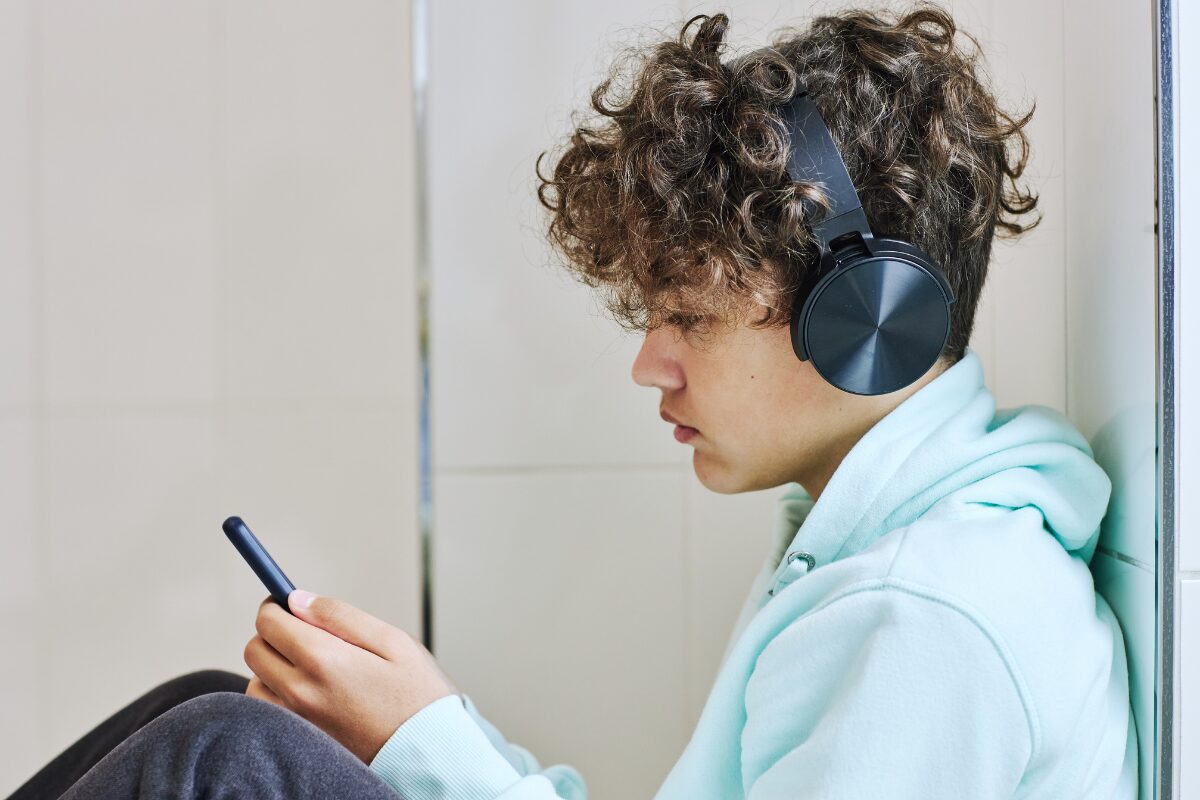 Teen sitting against a wall wearing headphones and a hoodie, looking at a smartphone.