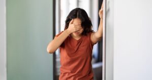 A young person stands in a hallway, leaning on the wall with one hand covering their eyes, showing fatigue and dizziness that can occur with POTS after COVID.