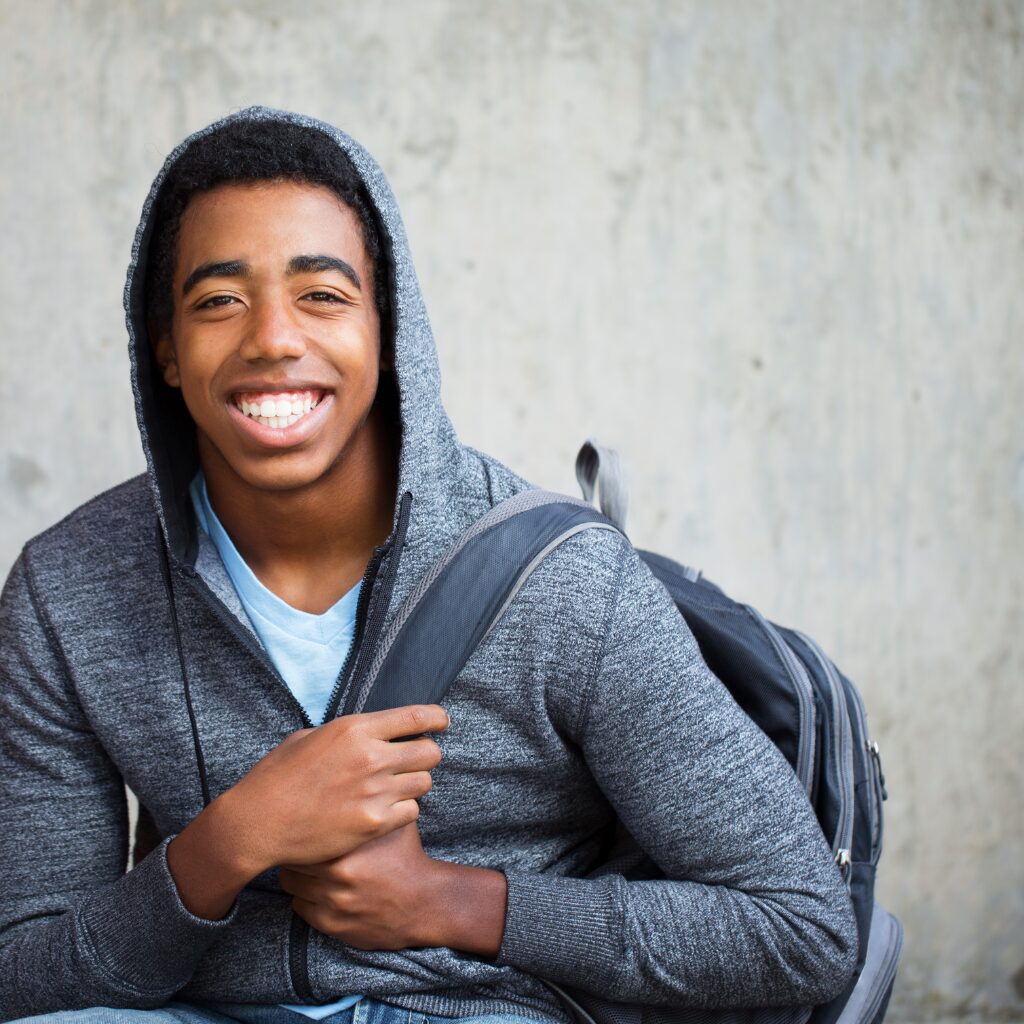 A smiling Black teenage boy wearing a gray hoodie and backpack sits against a concrete wall, holding one strap of his bag.