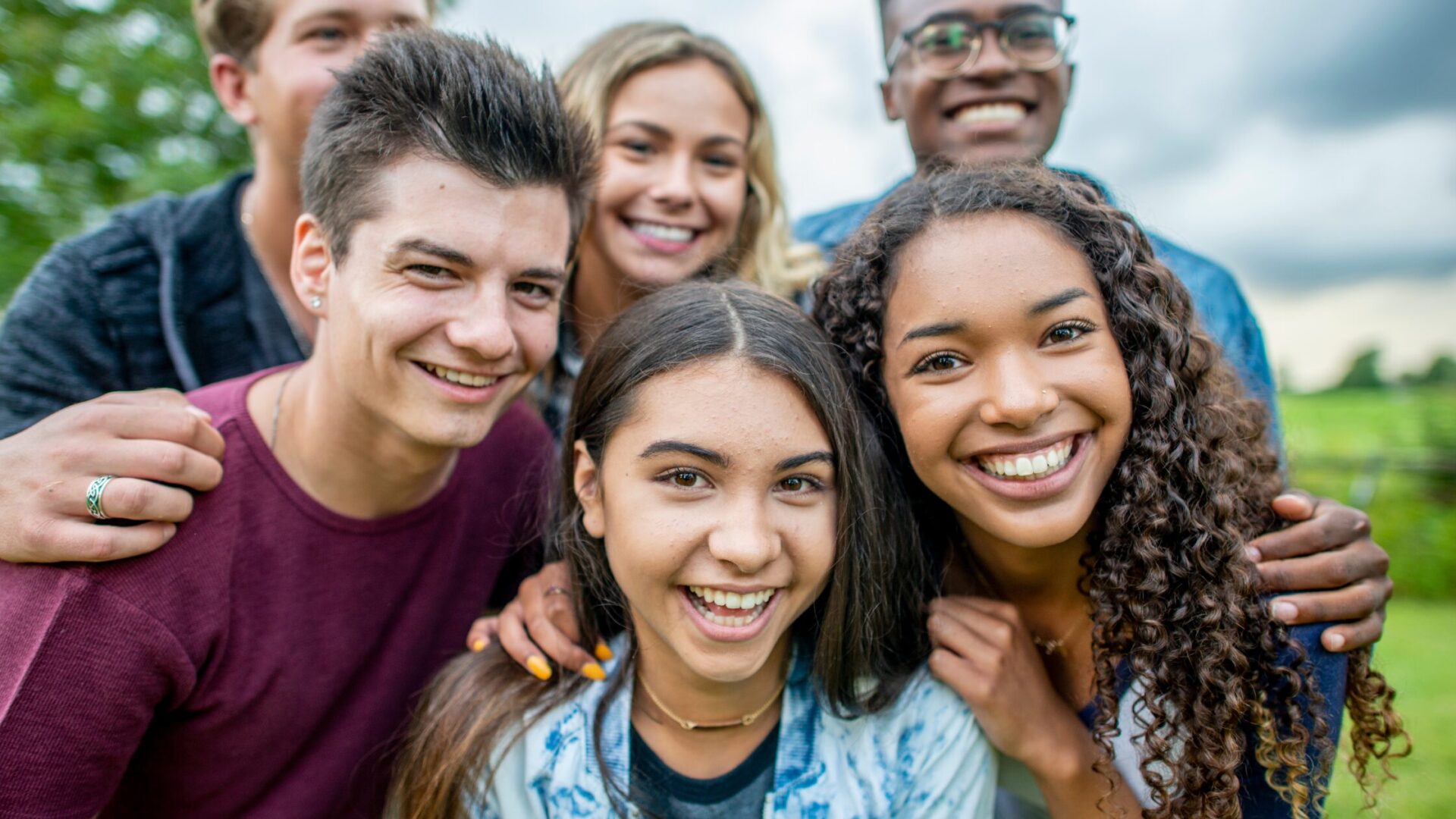 A group of smiling teens standing close together outdoors, showing friendship and energy on a cloudy day.