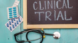 A chalkboard reading “Clinical Trial” beside blister packs of pills and a stethoscope on a blue wooden surface.