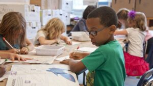 A school-age boy in glasses and a green shirt writes at a classroom table while other children work on art projects and worksheets around him.