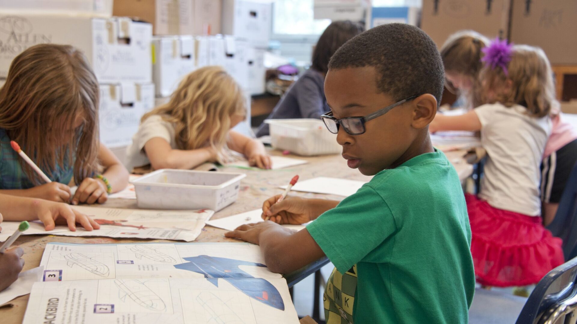 A school-age boy in glasses and a green shirt writes at a classroom table while other children work on art projects and worksheets around him.