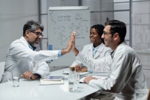 Colleagues in white lab coats gathered around a table, with two exchanging a high-five.