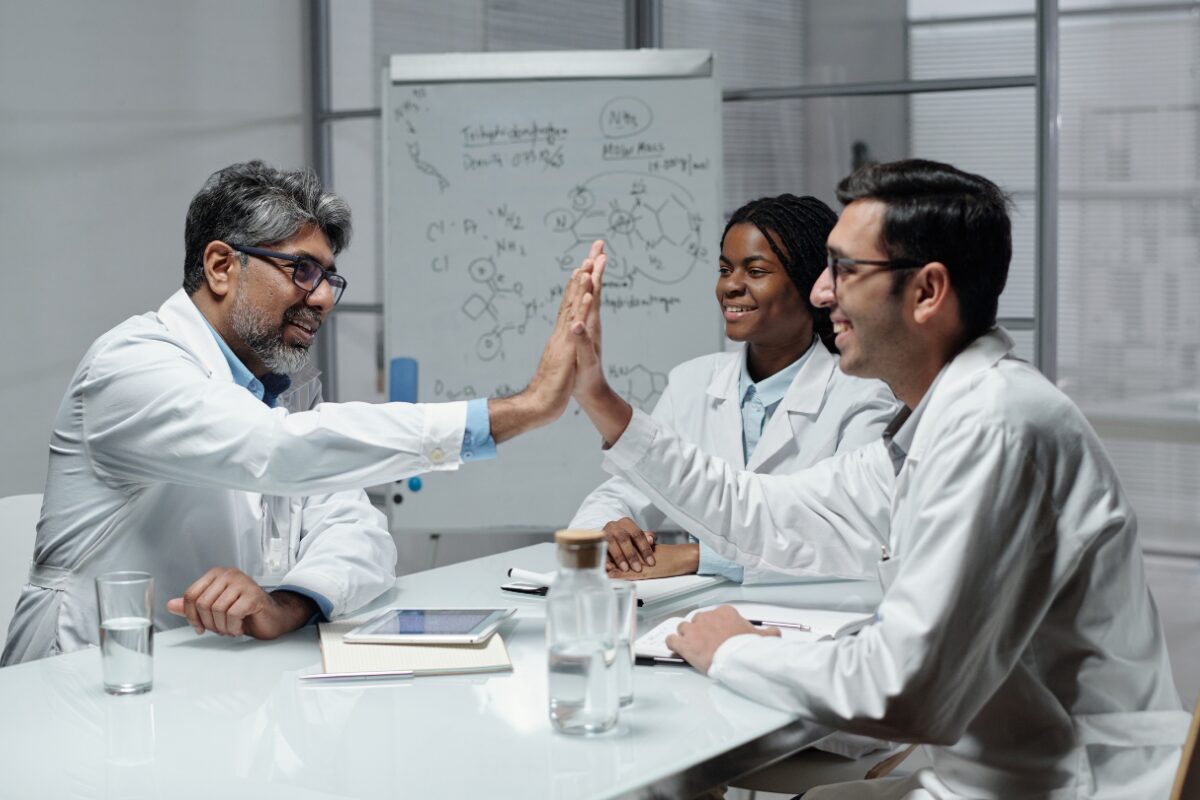 Colleagues in white lab coats gathered around a table, with two exchanging a high-five.