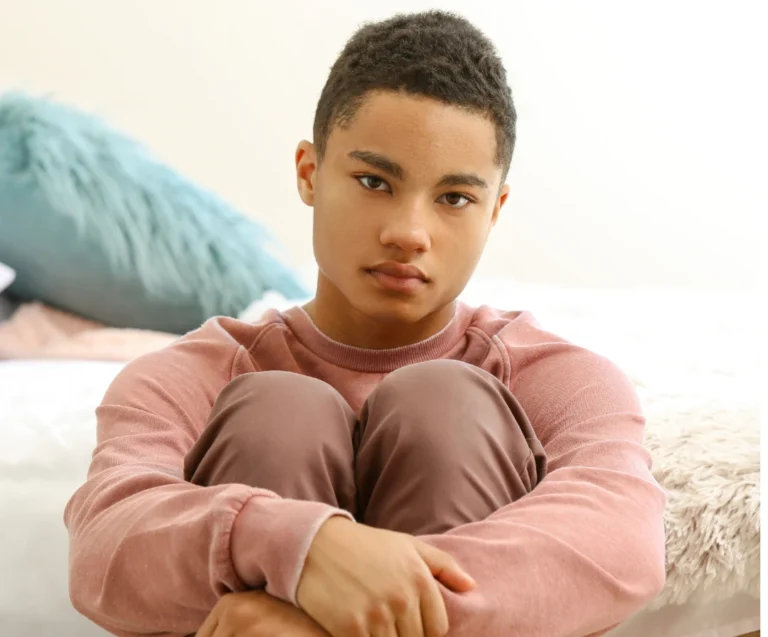 A young person sitting on a bed and hugging their knees.