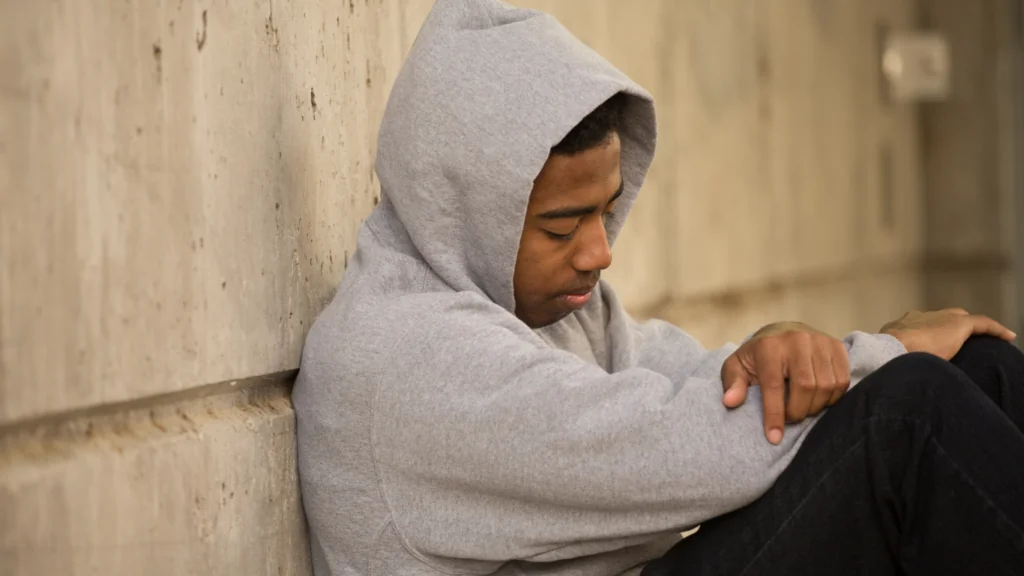 A young person in a gray hoodie sitting against a wall with arms over their knees and head lowered.