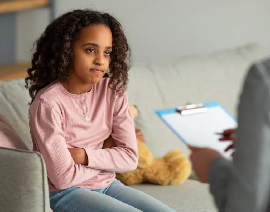 A child sitting on a couch and speaking with an adult holding a clipboard.
