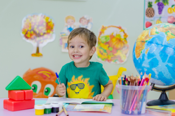 A child smiling at a desk with an open notebook, art supplies, and a globe.